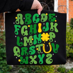 May include: Black tote bag featuring a green and yellow alphabet design with shamrocks, stars, and a horseshoe. The letters are in various shades of green with yellow accents. The bag is held up by a hand.