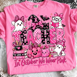 Op de afbeelding: Een felroze t-shirt met een grafisch ontwerp met het woord TEACHER in grote letters, omringd door illustraties van spoken, pompoenen, boeken en een heksenhoed, geaccentueerd met roze lintjes en tekst.