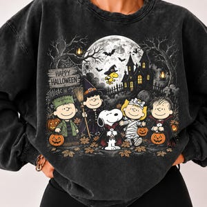 以下が含まれることがあります： ハロウィンの仮装をしたピーナッツのキャラクターのグラフィックプリントが施された、チャコールブラックのミネラルウォッシュ加工スウェットシャツ。デザインには、お化け屋敷、満月、コウモリ、そして木製の看板に書かれた「Happy Halloween」の文字が含まれています。