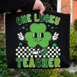 May include: Black tote bag featuring a cartoon clover with a smiling face, peace signs, and the text "ONE LUCKY TEACHER" in green. The clover is standing on a black and white checkered pattern. Small shamrocks and stars are scattered around the design.
