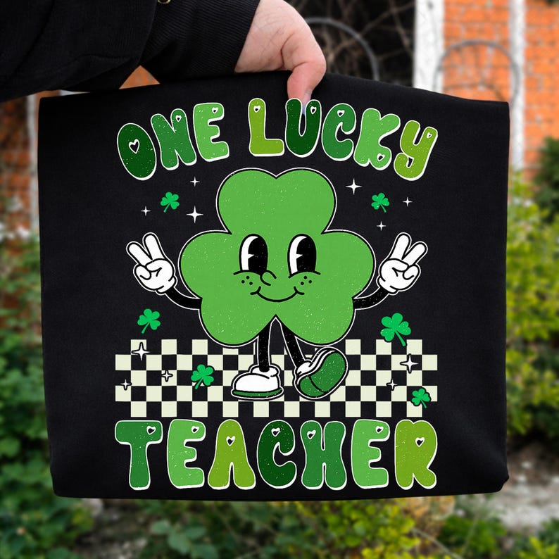 May include: Black tote bag featuring a cartoon clover with a smiling face, peace signs, and the text "ONE LUCKY TEACHER" in green. The clover is standing on a black and white checkered pattern. Small shamrocks and stars are scattered around the design.