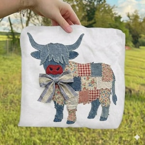 May include: A white square pillow featuring a patchwork design of a Highland cow. The cow has denim blue horns and shaggy hair, a red nose, and a bow. The body is made of various patterned fabrics.