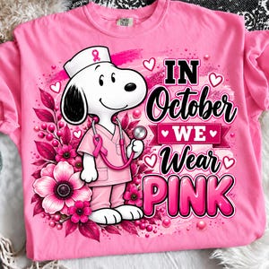 Puede incluir: Una camiseta rosa brillante con un gráfico de un perro de dibujos animados blanco vestido de enfermero con un estetoscopio y gorro rosas. El diseño incluye flores rosas, corazones y el texto "IN October WE WEAR PINK" en letras negras y rosas.