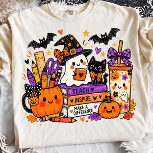 以下が含まれることがあります： ハロウィーンをテーマにしたカラフルなグラフィックがプリントされたクリーム色のTシャツ。デザインには、学用品が入ったカボチャのマグカップ、魔女の帽子をかぶったお化け、黒猫、TEACH INSPIRE MAKE A DIFFERENCEと書かれた本の山、紫色のリボンが付いたタンブラーが描かれています。