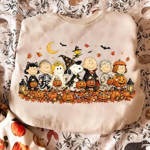 May include: A light beige crewneck sweatshirt featuring a colorful graphic of Peanuts characters dressed in Halloween costumes. The design includes Snoopy, Charlie Brown, and friends holding jack-o'-lanterns amidst autumn leaves, bats, and a crescent moon.