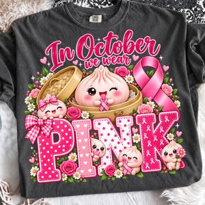Könnte beinhalten: Ein anthrazitfarbenes T-Shirt mit einem Aufdruck, der den Text In October we wear PINK zeigt. Das Design umfasst Cartoon-Teigtaschen in Bambusdämpfern, rosa Rosen, Gänseblümchen und eine große rosa Schleife zur Unterstützung des Bewusstseins.