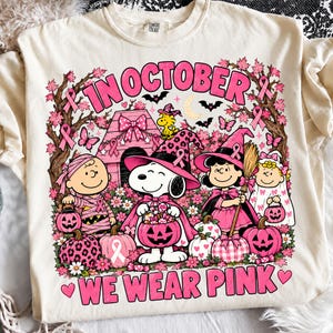 May include: A cream-coloured t-shirt featuring a graphic of Peanuts characters dressed in pink Halloween costumes. The text reads IN OCTOBER WE WEAR PINK. The design includes pink pumpkins, a doghouse, bats, and breast cancer awareness ribbons.