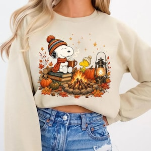 May include: A beige crewneck sweatshirt featuring a graphic print of Snoopy wearing a colorful knit hat and scarf, holding a mug of cocoa. The scene includes a campfire, a lantern, stacked books, pumpkins, and autumn leaves on a light background.