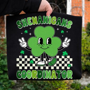 May include: Black tote bag with a green shamrock character giving a peace sign. The text "SHENANIGANS COORDINATOR" is in green and white. The shamrock has a smiling face and is wearing white shoes.