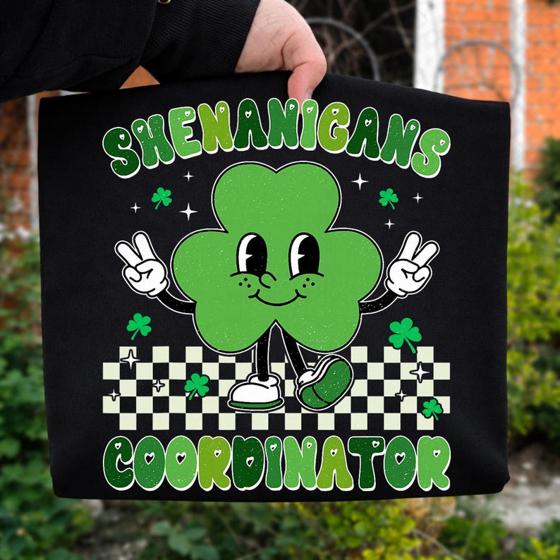 May include: Black tote bag with a green shamrock character giving a peace sign. The text "SHENANIGANS COORDINATOR" is in green and white. The shamrock has a smiling face and is wearing white shoes.