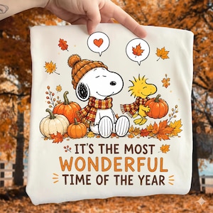 May include: A white t-shirt featuring a graphic of a white dog and a yellow bird wearing plaid scarves and hats, surrounded by orange pumpkins and autumn leaves. Above them are speech bubbles with a heart and a leaf. Text reads: IT'S THE MOST WONDERFUL TIME OF THE YEAR.