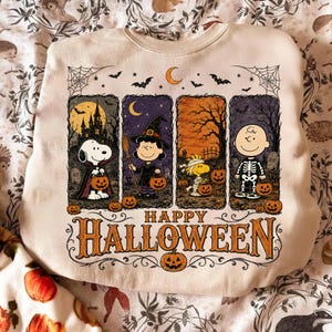以下が含まれることがあります： ハロウィーンの衣装を着たピーナッツのキャラクターのグラフィックプリントが施されたクリーム色のクルーネックトレーナー。スヌーピー、ルーシー、ウッドストック、チャーリー・ブラウンが描かれた4つのパネルと、その下にHappy Halloweenという文字がデザインされています。