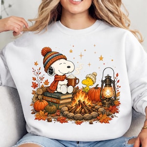 May include: A white sweatshirt featuring a graphic of Snoopy wearing a knit hat and scarf, holding a mug of cocoa while sitting on books. Beside Snoopy, Woodstock sits near a campfire, a lantern, and pumpkins, surrounded by autumn leaves.