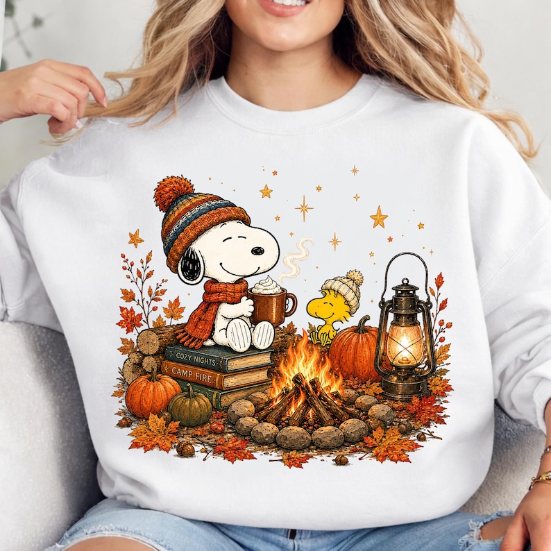 May include: A white sweatshirt featuring a graphic of Snoopy wearing a knit hat and scarf, holding a mug of cocoa while sitting on books. Beside Snoopy, Woodstock sits near a campfire, a lantern, and pumpkins, surrounded by autumn leaves.
