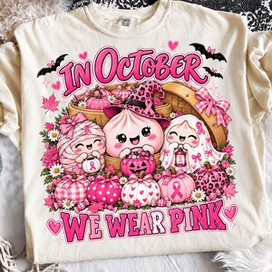 May include: A cream-coloured t-shirt featuring a graphic print with the text "IN OCTOBER WE WEAR PINK". The design includes three cute characters, pumpkins with various pink patterns, bats, and breast cancer awareness ribbons in a festive setting.