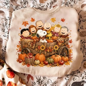 Pode incluir: Uma camisola de gola redonda cor de creme com uma impressão gráfica das personagens Peanuts sentadas num carro de madeira cheio de abóboras, girassóis e folhas de outono. O design inclui o Snoopy, o Charlie Brown e amigos.