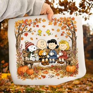 Puede incluir: Un cuadrado de tela blanco con una ilustración de Peanuts de Snoopy, Charlie Brown, Lucy y Sally sentados en un banco de madera bajo árboles otoñales. Sostienen tazas mientras dos pájaros amarillos vuelan encima. Calabazas y hojas rodean la escena.