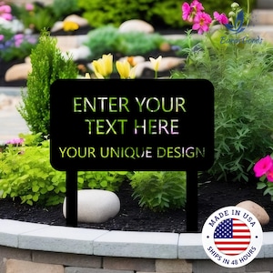May include: Black metal garden sign with the text "ENTER YOUR TEXT HERE YOUR UNIQUE DESIGN" in a cut-out style. The sign is on two posts and set in a garden bed with flowers and greenery. A "Made in USA" sticker is visible.