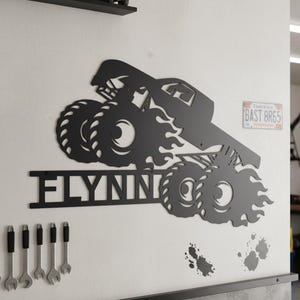 May include: Black metal wall art featuring a monster truck design with the name "FLYNNIN" cut out below. The truck has large tires and flames. A license plate is visible in the background.