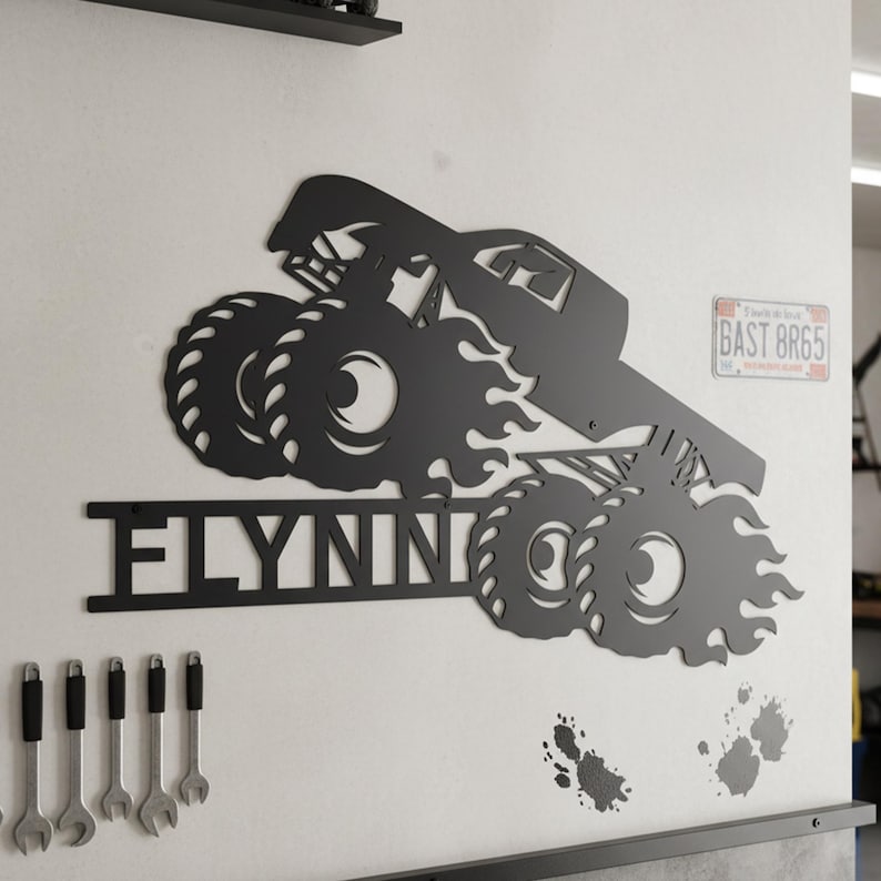 May include: Black metal wall art featuring a monster truck design with the name "FLYNNIN" cut out below. The truck has large tires and flames. A license plate is visible in the background.