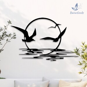 May include: Black metal wall art depicting three seagulls in flight around a circular sun, above a stylized water line. The artwork is mounted on a white wall, with the BuracGoods logo in the upper right corner.