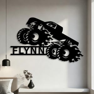 May include: Black metal wall art featuring a monster truck design with flames and the name "FLYNN" cut out below. The truck has large tires and is angled as if in motion. This decorative piece is suitable for a bedroom or living room.