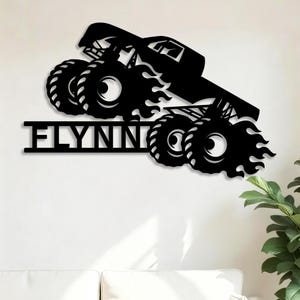 May include: Black metal wall art featuring a monster truck design with large tires and flames. The word "FLYNN" is displayed in bold letters below the truck. The artwork is mounted on a white wall.