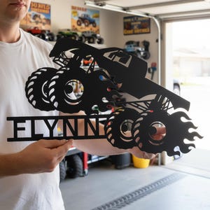 May include: Black metal monster truck sign with the name "FLYNN" cut out below the truck. The truck has large tires and flames. The sign is held up in front of a garage.