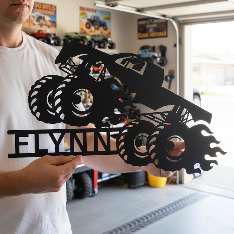 May include: Black metal monster truck sign with the name "FLYNN" cut out below the truck. The truck has large tires and flames. The sign is held up in front of a garage.