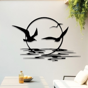 May include: Black metal wall art depicting three birds in flight against a circular sun or moon. Below, stylized lines represent water. The artwork is mounted on a white wall, creating a striking contrast. A decorative piece for home decor.