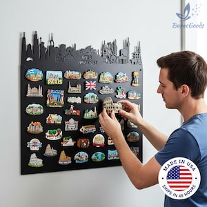 May include: A black metal display board with a skyline silhouette and the words "OUR JOURNEY". The board holds numerous colorful souvenir magnets of landmarks from around the world. A person is holding a magnet of the Colosseum.