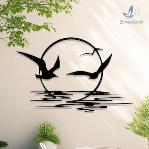 May include: Black metal wall art depicting three seagulls in flight within a circular frame, above stylized water lines. The design includes the text "BuracGoods" in the upper right corner. The artwork is mounted on a light-colored wall.