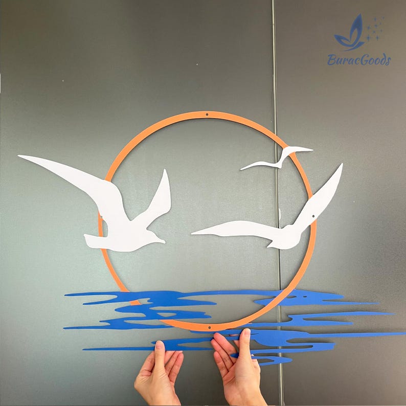May include: A wall art piece featuring a sunset scene with white seagulls, an orange sun, and blue water. The artwork is made of cut-out shapes and is designed to be mounted on a wall. The brand name "BuracGoods" is visible.