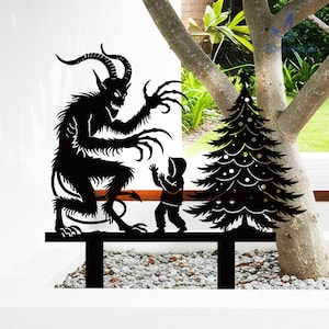 May include: Black metal yard art depicting a Krampus figure menacing a child near a Christmas tree. The silhouette shows the horned beast with long claws and a child in a hooded coat. The Christmas tree has decorative cutouts.