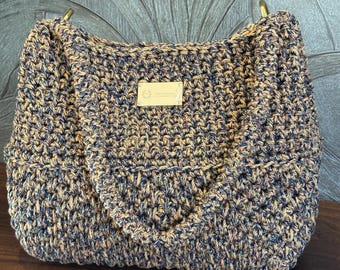 Könnte beinhalten: Eine braune und schwarze gehäkelte Tragetasche mit goldenen Metallgriffen. Die Tasche hat ein kleines braunes Lederetikett mit dem Wort "Handmade" darauf.