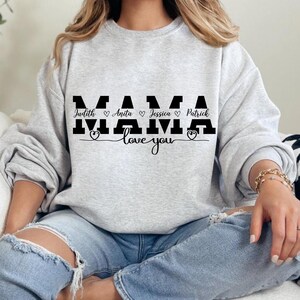 Könnte beinhalten: Grauer Pullover mit dem Wort "MAMA" in schwarzen Buchstaben. Die Namen Judith, Anita, Jessica und Patrick sind in kleineren Buchstaben unter dem Wort "MAMA" mit einem Herzsymbol zwischen jedem Namen geschrieben. Der Text "love you" ist unter den Namen mit einem Herzsymbol am Anfang und Ende des Textes geschrieben.