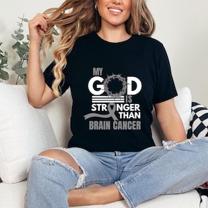 Könnte beinhalten: Schwarzes T-Shirt mit einem weißen Aufdruck, der "My God is Stronger Than Brain Cancer" lautet, mit einem grauen Band und einer Dornenkrone.