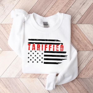 Könnte beinhalten: Weißer Pullover mit einem abgenutzten amerikanischen Flaggenmuster und dem Wort "TARIFFED" in roten Buchstaben.