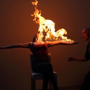 May include: A person seated with arms outstretched, appearing to be performing a fire-breathing act. Flames erupt from the person's mouth, creating a dramatic visual. Another person is in the background, seemingly part of the performance.