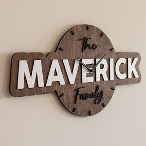 May include: A wooden wall clock with the text "The Maverick Family" in white and dark brown. The clock hands are black. The clock is mounted on a cream wall.