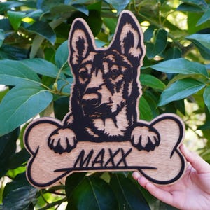 May include: Wooden dog-themed sign featuring a detailed black and brown German Shepherd design. The dog is holding a bone-shaped sign with the name "MAXX" in a stylish font. The sign is made of wood and is set against a backdrop of green leaves.