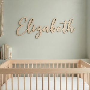 Può includere: Una scritta da parete in legno naturale con il nome Elizabeth in un carattere corsivo. La scritta è montata su una parete sopra una culla in legno chiaro. Il testo Font: 12 Color: Natural è visibile nell'angolo in basso a destra dell'immagine.