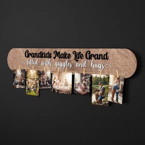 May include: Wooden photo display with the text "Grandkids Make Life Grand filled with giggles and hugs." The display holds several photos clipped with small wooden clothespins. The wooden board has a light brown finish and is mounted on a dark background.