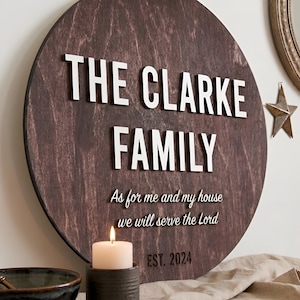 May include: A circular dark brown wooden sign featuring white raised lettering that reads THE CLARKE FAMILY, followed by the script text As for me and my house we will serve the Lord, and the text EST. 2024 at the bottom of the wooden board.