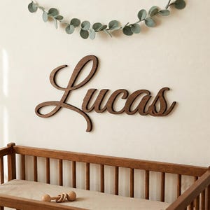 May include: A dark brown wooden wall sign featuring the name Lucas in a cursive script font. The sign is mounted on a plain wall above a wooden crib, with a decorative garland of green leaves hanging horizontally above the name.
