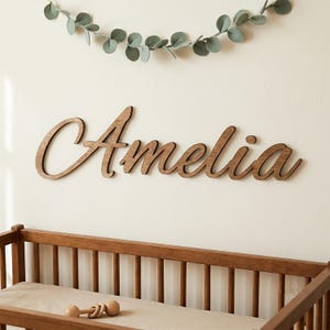 May include: A wooden wall sign featuring the name Amelia in a cursive script font. The letters are dark brown with a smooth finish. Below the sign is a wooden crib with a light-coloured mattress and a small wooden rattle toy resting on it.