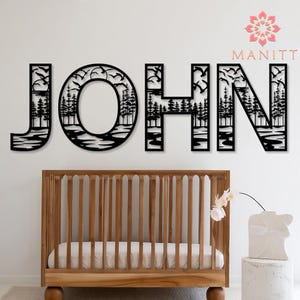 Pode incluir: Arte de parede em metal preto com a palavra "JOHN", apresentando uma cena da natureza com &aacute;rvores e p&aacute;ssaros. As letras s&atilde;o montadas acima de um ber&ccedil;o de madeira com um colch&atilde;o branco. Um vaso branco com uma flor est&aacute; em um pequeno pedestal.