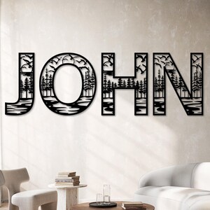 Pode incluir: Arte de parede em metal preto com o nome "JOHN". As letras s&atilde;o estilizadas com uma cena da natureza, incluindo &aacute;rvores, &aacute;gua e p&aacute;ssaros voando. A obra de arte &eacute; montada em uma parede de cor neutra em uma sala de estar.