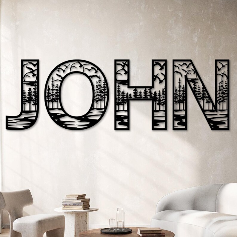 Pode incluir: Arte de parede em metal preto com o nome "JOHN". As letras s&atilde;o estilizadas com uma cena da natureza, incluindo &aacute;rvores, &aacute;gua e p&aacute;ssaros voando. A obra de arte &eacute; montada em uma parede de cor neutra em uma sala de estar.