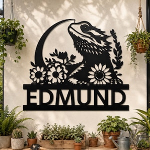 May include: A black metal wall sign featuring a silhouette of a bearded dragon lizard positioned in front of a crescent moon and floral elements. The name EDMUND is cut out in bold capital letters on a horizontal bar at the bottom of the design.